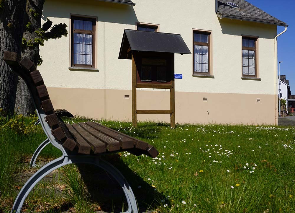 Ein Holzbänkchen mit Metallgestell steht auf einer Wiese mit Gänseblümchen vor einem hellen Haus mit einem kleinen Holzhäuschen – typisch für den naturnahen Charakter von Charlottenberg im Rhein-Lahn-Kreis.