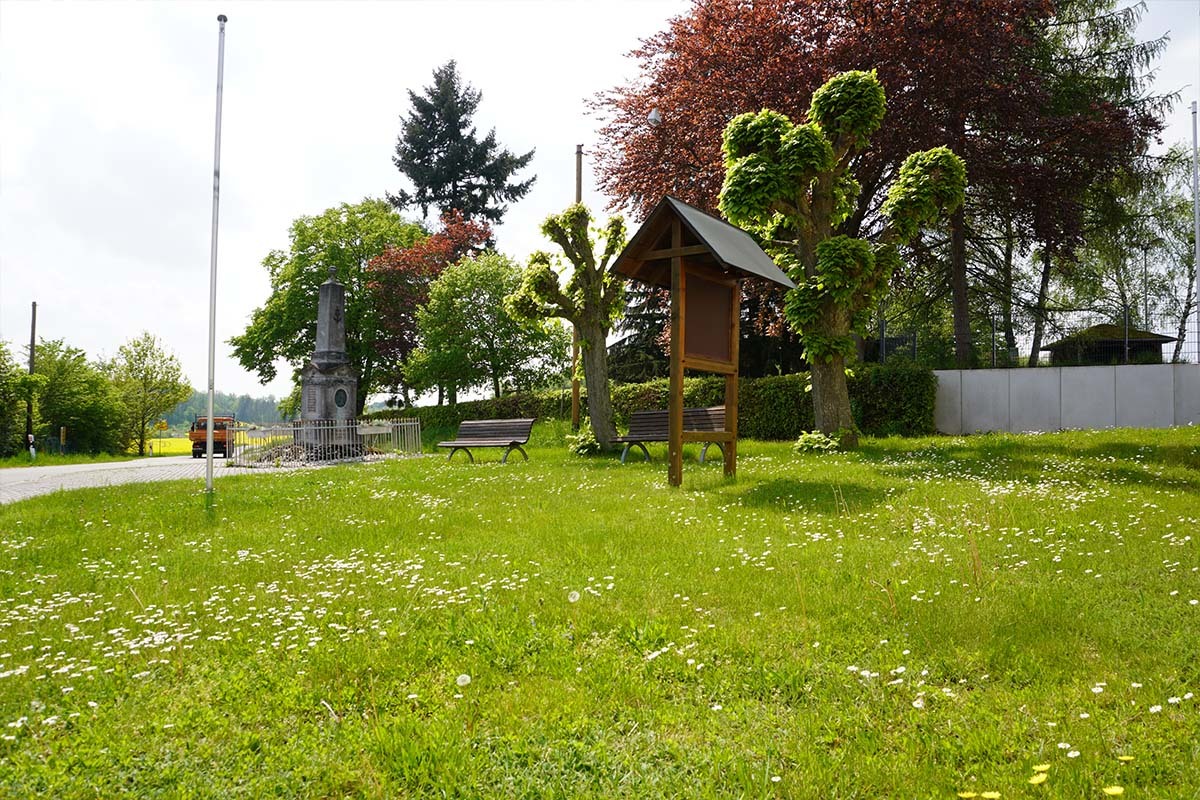 Grüne Wiese mit Gänseblümchen, einem Denkmal, Bänken und einem Holzschildhäuschen in Charlottenberg – ein ruhiges, naturnahes Gemeindesetting mit Bäumen und Freizeitmöglichkeiten.