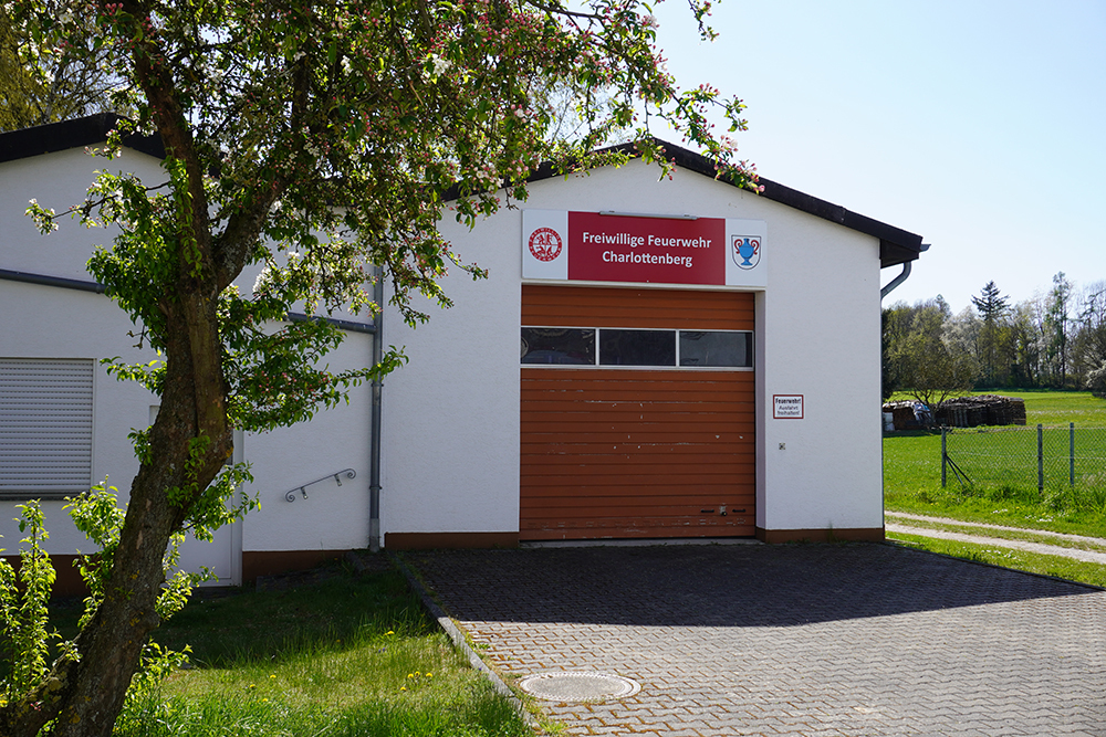 Gebäude der Freiwilligen Feuerwehr Charlottenberg mit roter Beschriftung, braunem Garagentor und umgebendem Grün in ländlicher Umgebung.