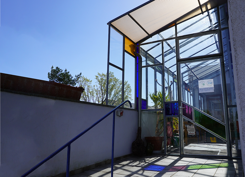 Modernes Glas-Eingangstor mit farbigen Paneelelementen, blauer Handlauf und Pflanzenbeeten vor einem öffentlichen Gebäude in Charlottenberg, Rhein-Lahn-Kreis, unter klarem Himmel.