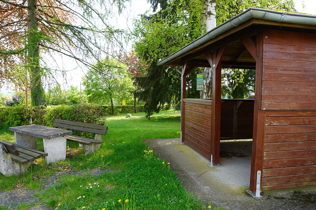 Holzunterstand mit Picknicktisch und Bänken in einem grünen, naturnahen Freizeitbereich der Ortsgemeinde Charlottenberg, umgeben von Bäumen und Gras.