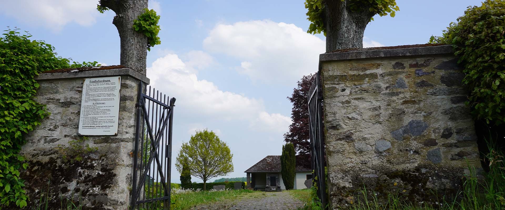 Eingang zum Friedhof der Ortsgemeinde Charlottenberg: steinernes Tor mit Eisengitter, Schild 