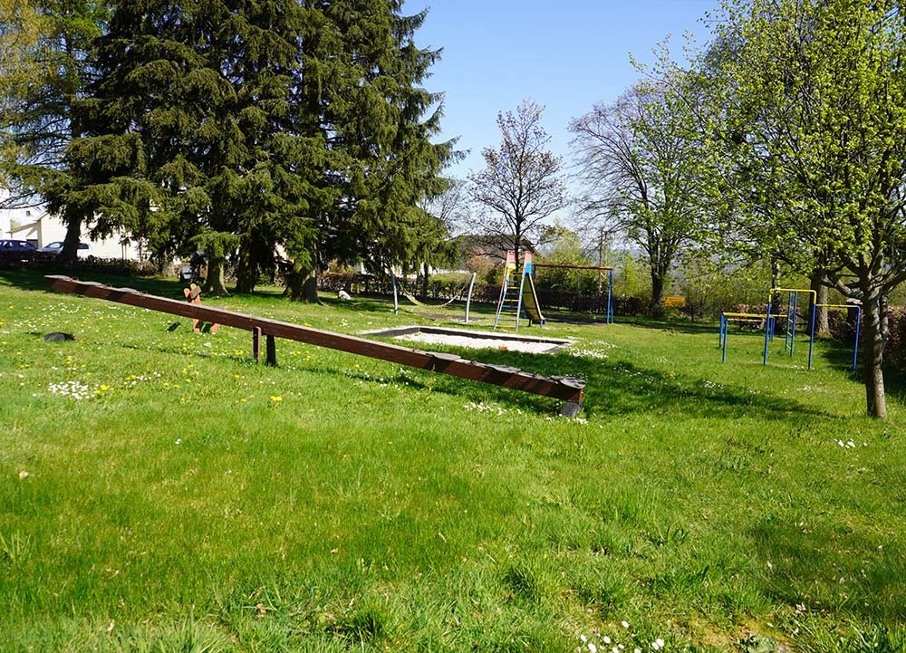 Spielplatz in Charlottenberg mit Wippe, Klettergeräten und Bäumen auf grünem Rasen im sonnigen Naturpark Nassau.