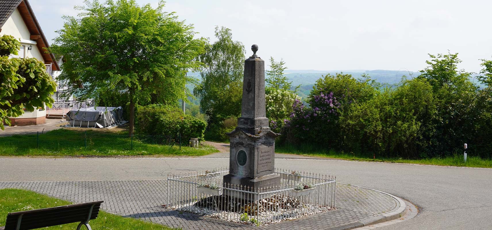 Ein historisches Denkmal mit Metallzaun auf dem Dorfplatz von Charlottenberg, umgeben von Bäumen, Büschen und einem Haus, mit Blick auf die Hügel des Naturparks Nassau im Hintergrund.
