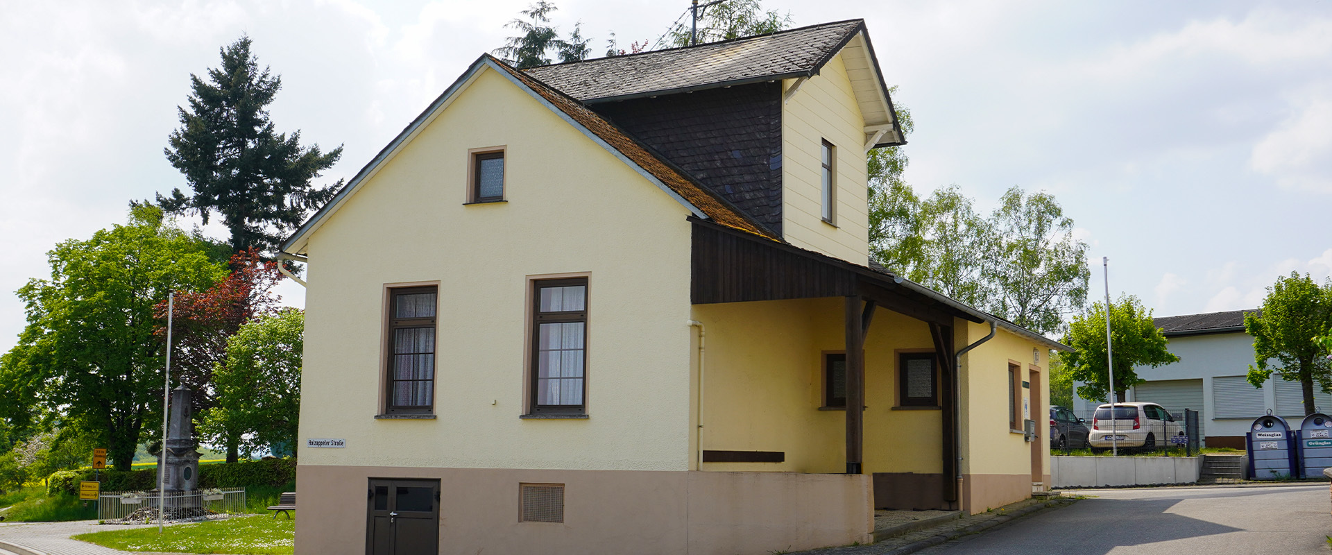 Das zweistöckige Dorfgemeinschaftshaus in Charlottenberg mit gelber Fassade, dunklen Holzelementen und Satteldach steht im Dorfzentrum, umgeben von Bäumen und Grünflächen – ein zentraler Treffpunkt für die Gemeinde.