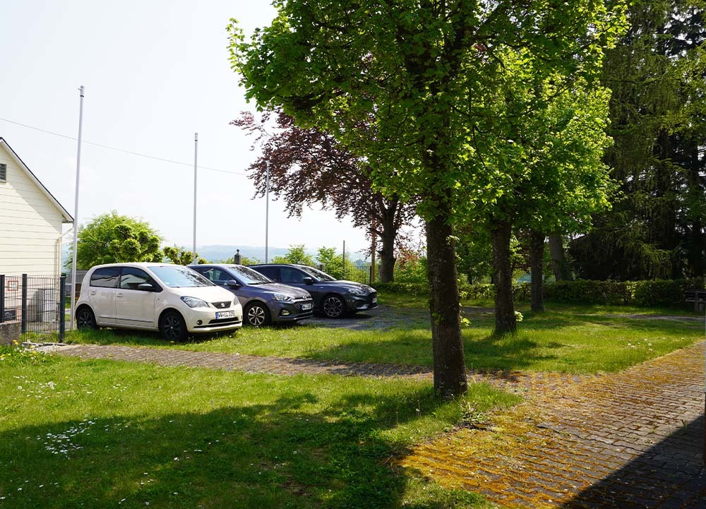 Parkplatz mit drei Autos (weiß, grau, dunkel) auf grasigem Gelände mit Bäumen und Pflasterweg – typisch für den naturnahen Ort Charlottenberg im Naturpark Nassau.