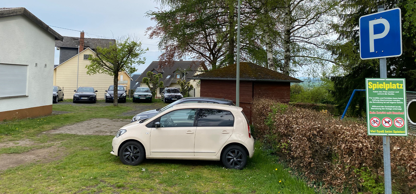 Parkplatz mit Spielplatzschild in Charlottenberg: Ein hellbeiges Auto steht auf dem Gras neben einem blauen 'P'-Schild und einem grünen 'Spielplatz'-Schild, umgeben von Häusern, Bäumen und Büschen – typisch für die naturnahe Ortsgemeinde im Rhein-Lahn-Kreis.