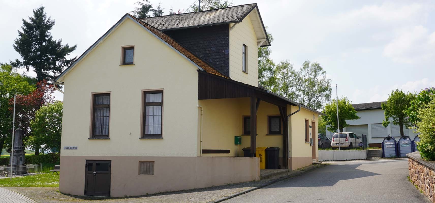 Ein zweistöckiges Gebäude mit gelber Fassade und dunklem Dach, vermutlich eine kommunale Einrichtung in Charlottenberg, steht am Straßenrand mit Bäumen und Nachbargebäuden im Hintergrund.