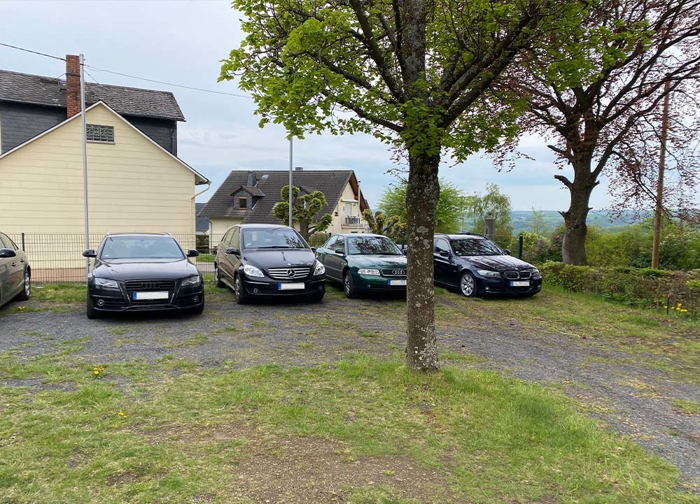 Parkplatz mit mehreren Autos vor Wohnhäusern und Bäumen in Charlottenberg, einem naturnahen Ort am Rand des Naturparks Nassau.