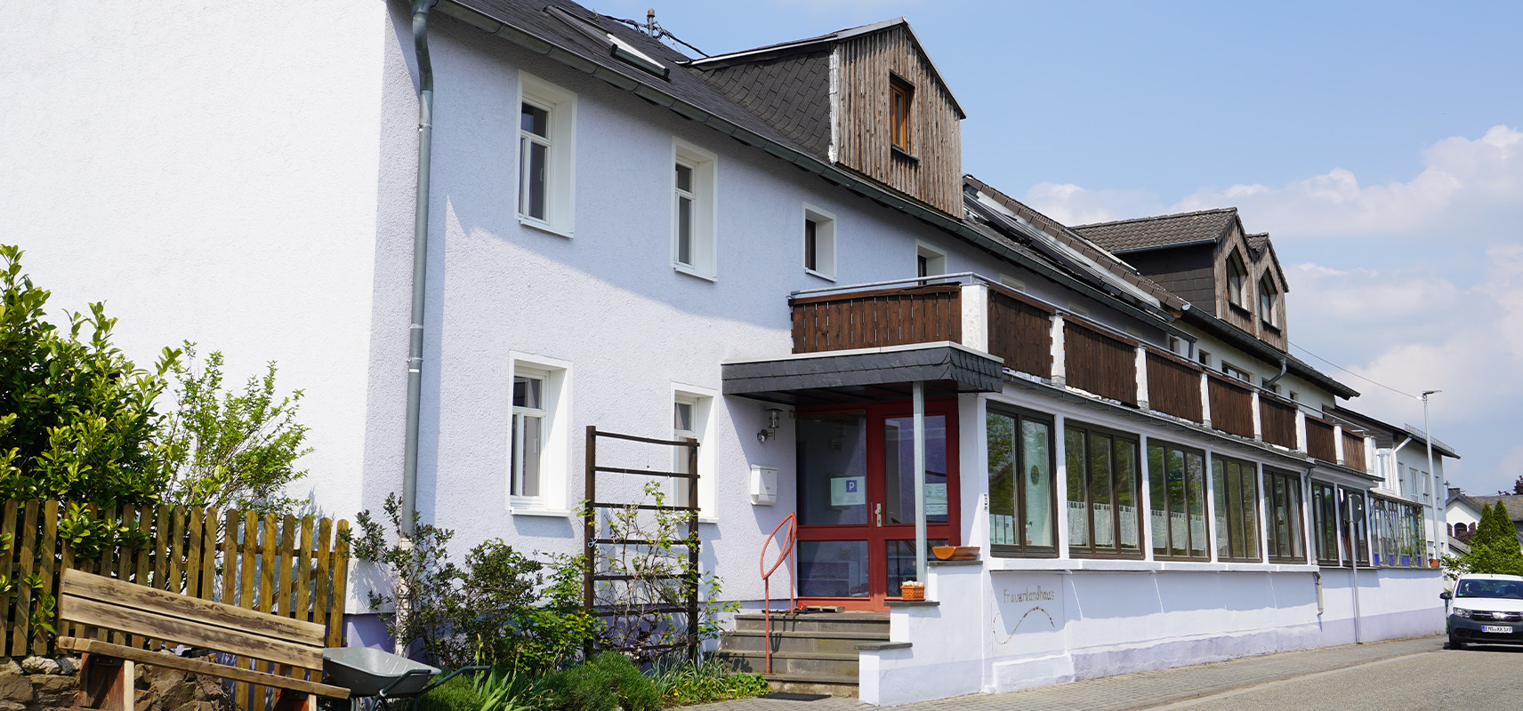 Traditionelles hellblaues Gebäude mit roter Tür und Holzveranda in Charlottenberg (Rhein-Lahn-Kreis), das als lokale Einrichtung oder Gemeinschaftshaus dient.